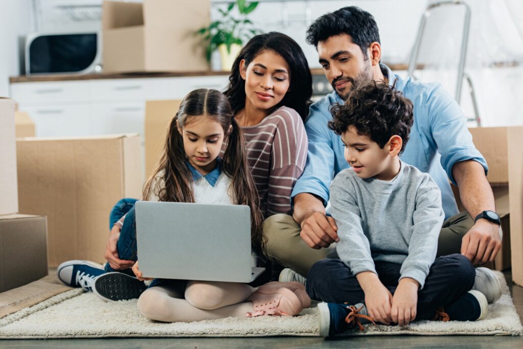 cheerful latino family looking at laptop while sitting on carpet in new home
